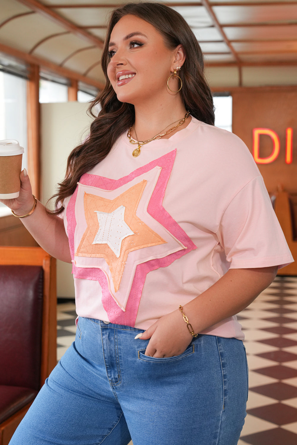 Woman wearing a pink t-shirt with a colorful star design, holding a drink in a casual setting.